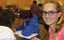 A girl on the right smiles as other class members in the background are studying or doing science experiements